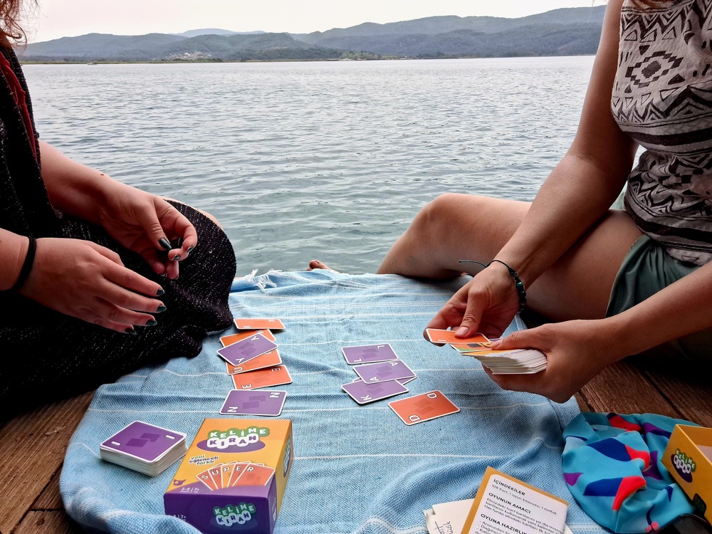 Players sitting on a jetty by the water, hands over a game of Kelime Kıran